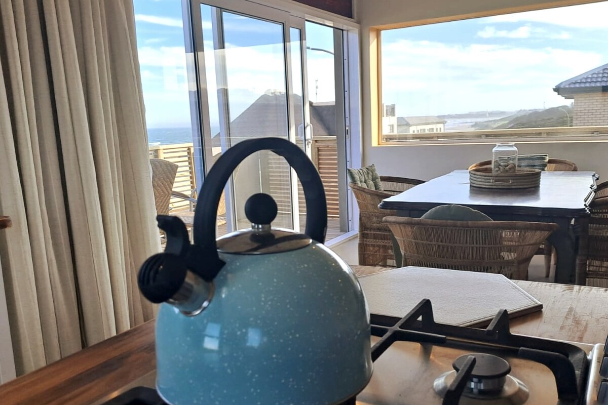 A kitchen space is highlighted by a light blue kettle on a stove. A dining area with natural wood accents is visible, featuring a large table and wicker chairs. Large glass doors allow ample natural light and offer views of the surrounding landscape.