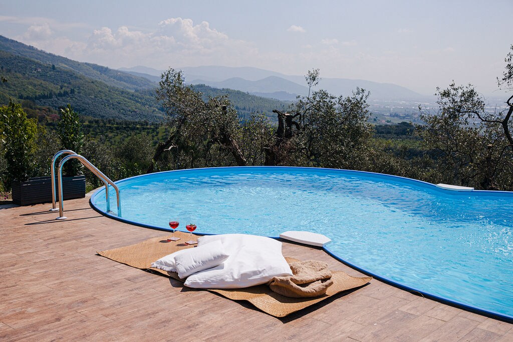 A turquoise pool is set against a backdrop of rolling hills and olive trees, with gentle waves lapping at the edge. A cozy blanket and cushions are arranged on a sunlit patio, inviting relaxation. Two glasses are placed nearby, suggesting a leisurely afternoon.