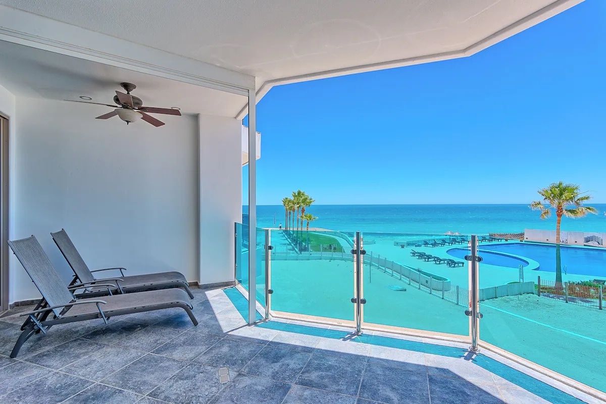 A spacious balcony features glass railings overlooking a serene ocean view. Two lounge chairs are positioned for relaxation, while a ceiling fan adds comfort. The background reveals a pool area and palm trees, enhancing the coastal experience.