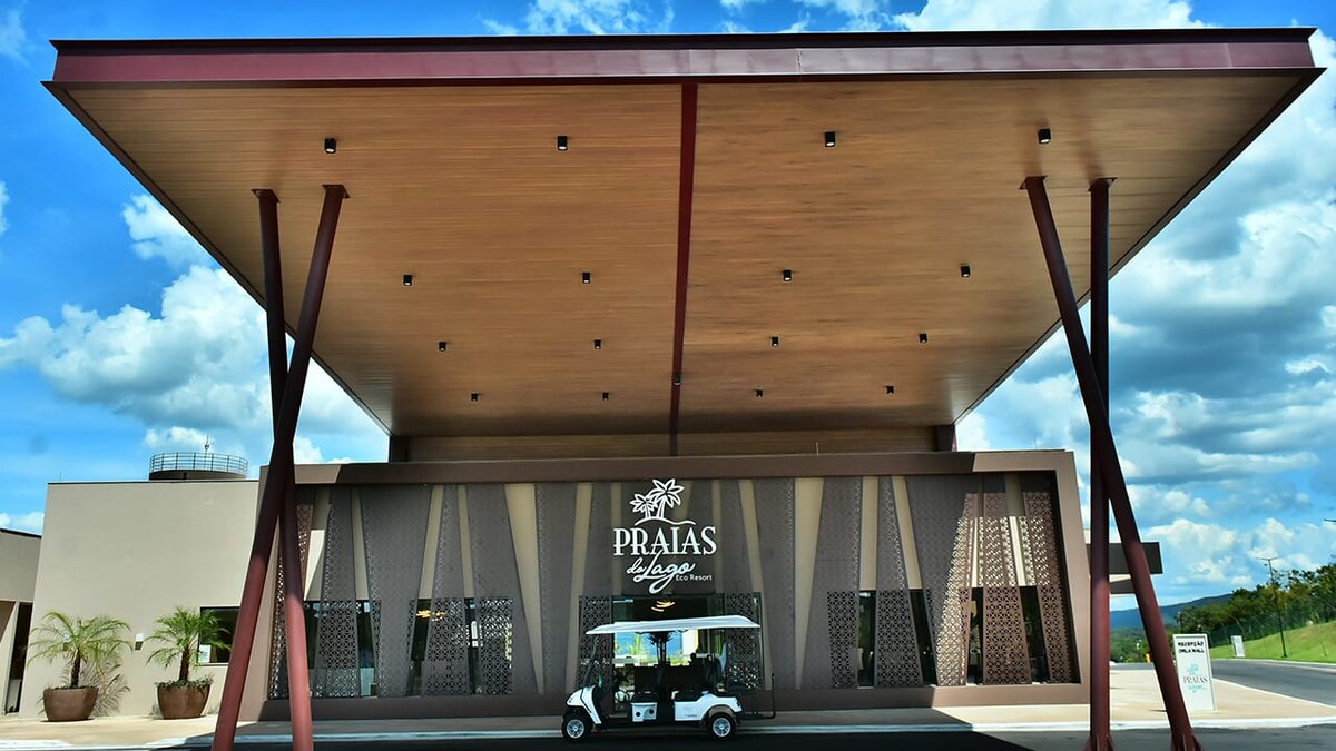 The entrance of Praias Lodge is showcased with a large, modern roof supported by tall beams. The facade features intricate patterned screens, providing a glimpse of the inviting lobby area. A golf cart is parked at the front, welcoming guests to explore the surroundings.