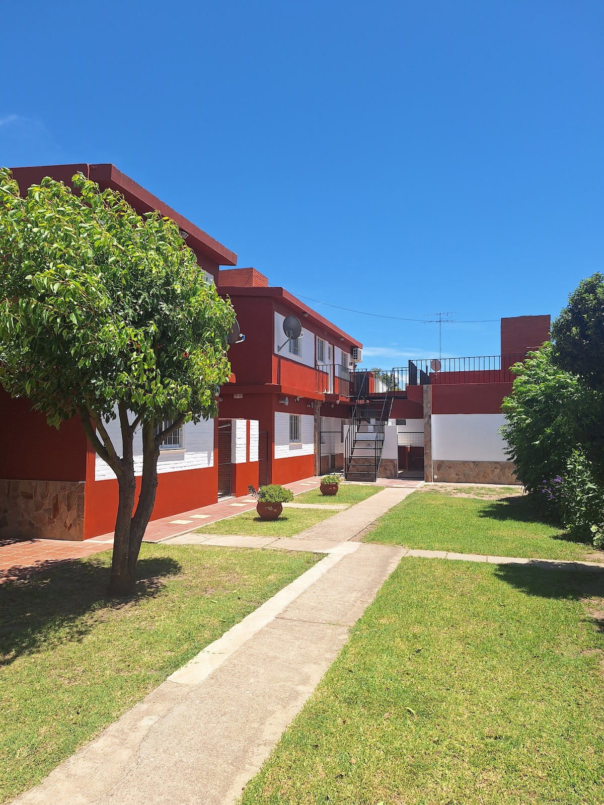 The exterior of a red and white building complex is emphasized, featuring neatly landscaped green grass and small trees. Pathways lead through the courtyard, connecting multiple units while ensuring easy access to the surrounding area under a clear blue sky.