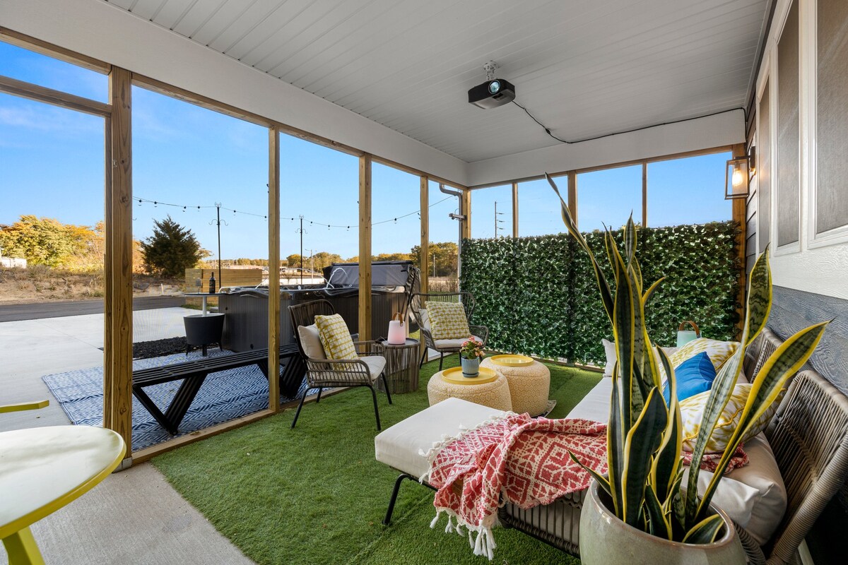 An outdoor screened-in porch features comfortable seating arrangements with colorful cushions and a large coffee table. A green lawn-like carpet covers the floor. A projector is mounted on the ceiling, while a hot tub is visible beyond the porch, surrounded by greenery.