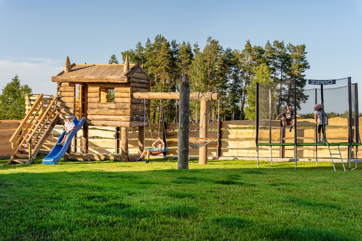A wooden play structure is set against a backdrop of trees, featuring a slide, swings, and climbing elements. Nearby, a large trampoline offers additional play options. A lush green lawn surrounds the play area, providing a spacious environment for outdoor activities.