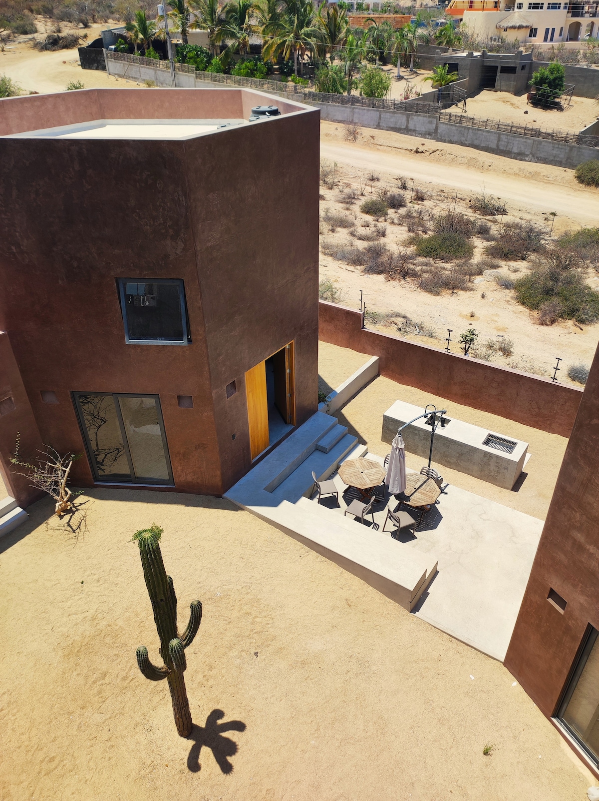 An aerial view of a minimalist casita surrounded by desert landscape features a small table with chairs and a shaded umbrella on a concrete patio. A tall cactus casts a long shadow, enhancing the tranquil outdoor space.