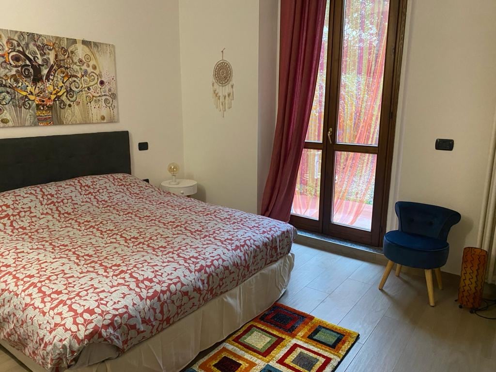 A cozy sleeping area features a spacious bed covered with a patterned duvet, complemented by a colorful area rug beneath. A small blue chair and a decorative lamp are placed nearby. Large windows, adorned with curtains, allow natural light to illuminate the room.