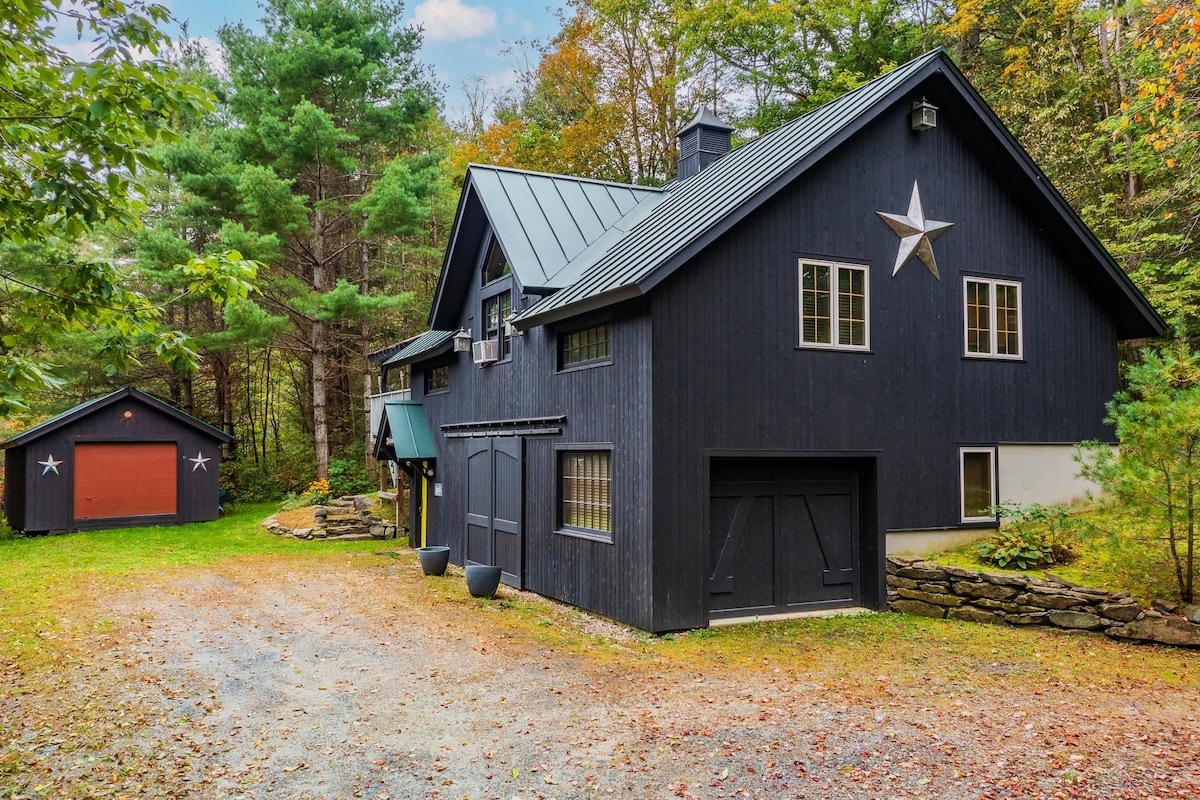 A charming black house with a distinctive star decoration stands amidst a lush green landscape. The property features a gravel driveway leading to the front entrance, with trees framing the surroundings and a second structure visible on the left.
