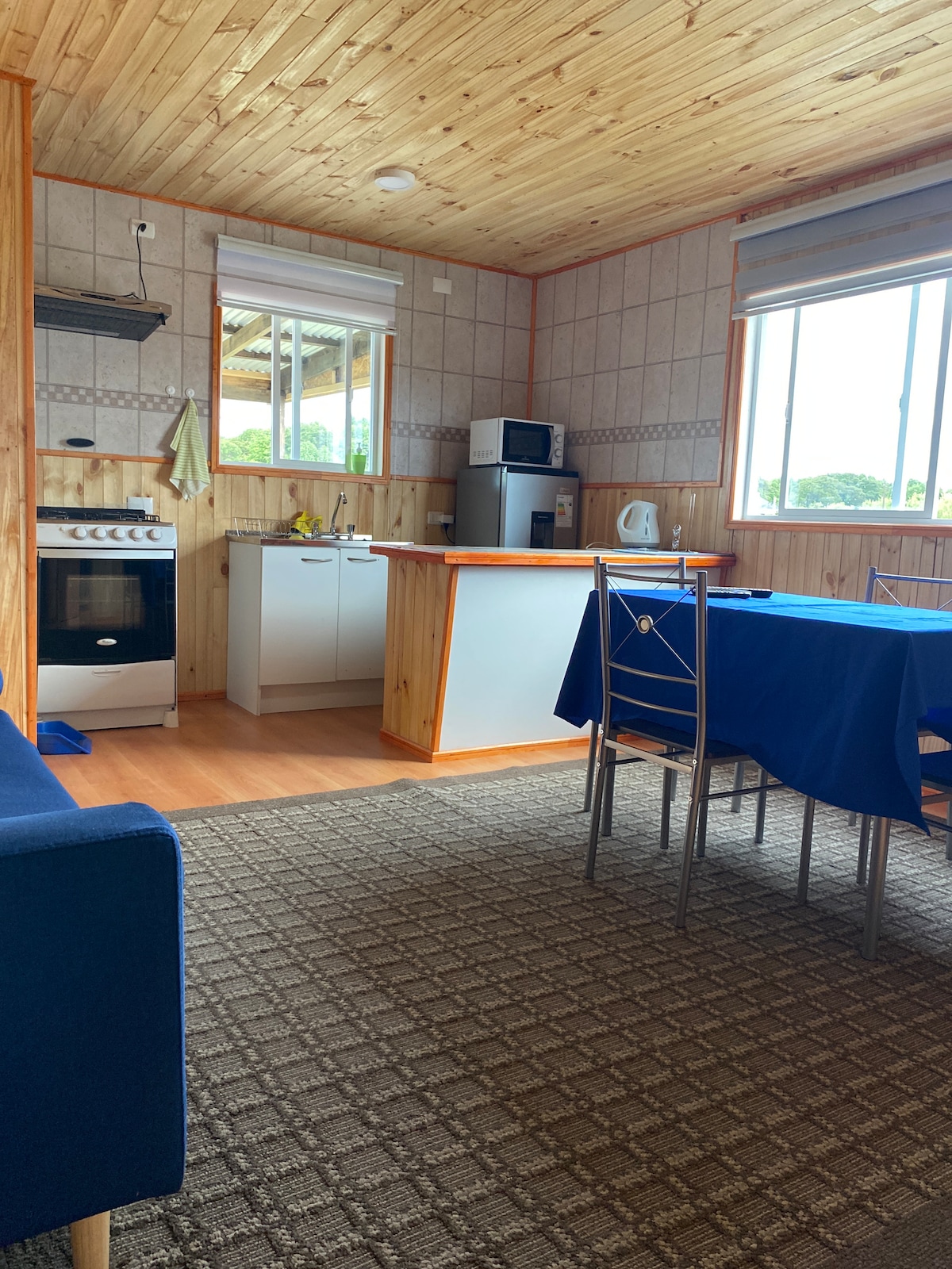A cozy kitchen space features wooden walls and a sloped ceiling. An oven, microwave, and refrigerator are visible along with a small countertop. A blue tablecloth covers a table surrounded by metal chairs. Natural light enters through a large window, illuminating the warm interior.