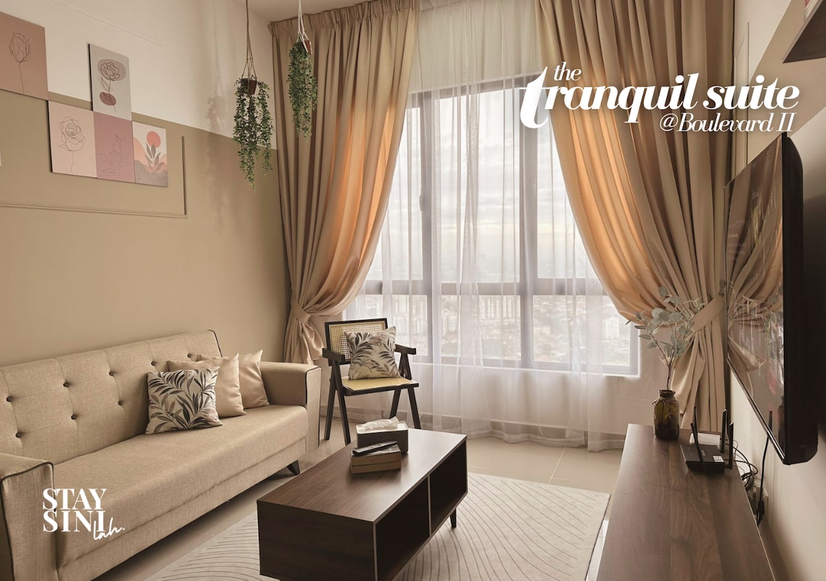 A bright living area features large windows with sheer curtains allowing natural light. A cream-colored sofa is positioned against the wall, accompanied by a wooden armchair. A coffee table sits in front, while decorative plants and framed artwork enhance the space.