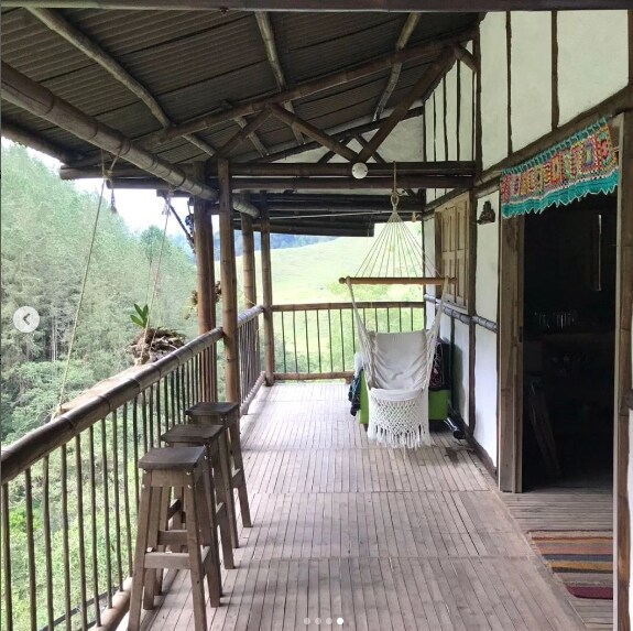A spacious balcony is framed by bamboo railings, featuring two wooden stools and a hammock for relaxation. The wooden floor enhances the rustic charm. Natural greenery is visible in the background, creating a serene environment for enjoying the outdoors.