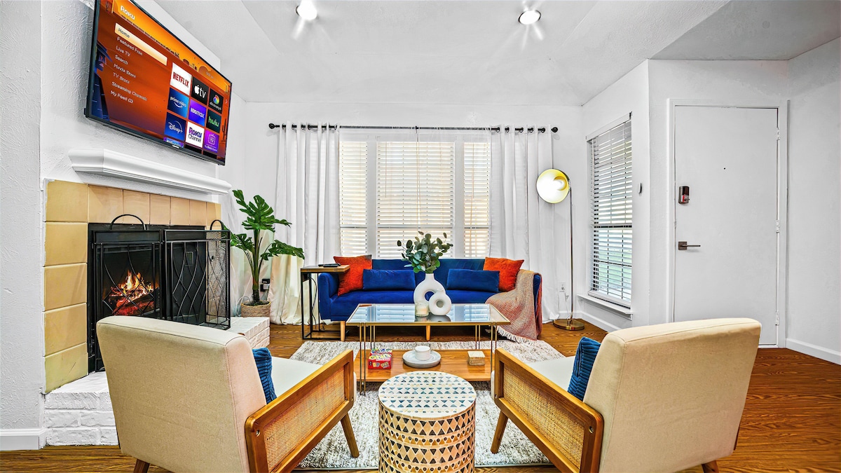 A modern living area features a fireplace with a television mounted above. Comfortable seating is arranged around a coffee table, complemented by decorative pillows. Large windows allow natural light to fill the space, while houseplants add a touch of greenery.