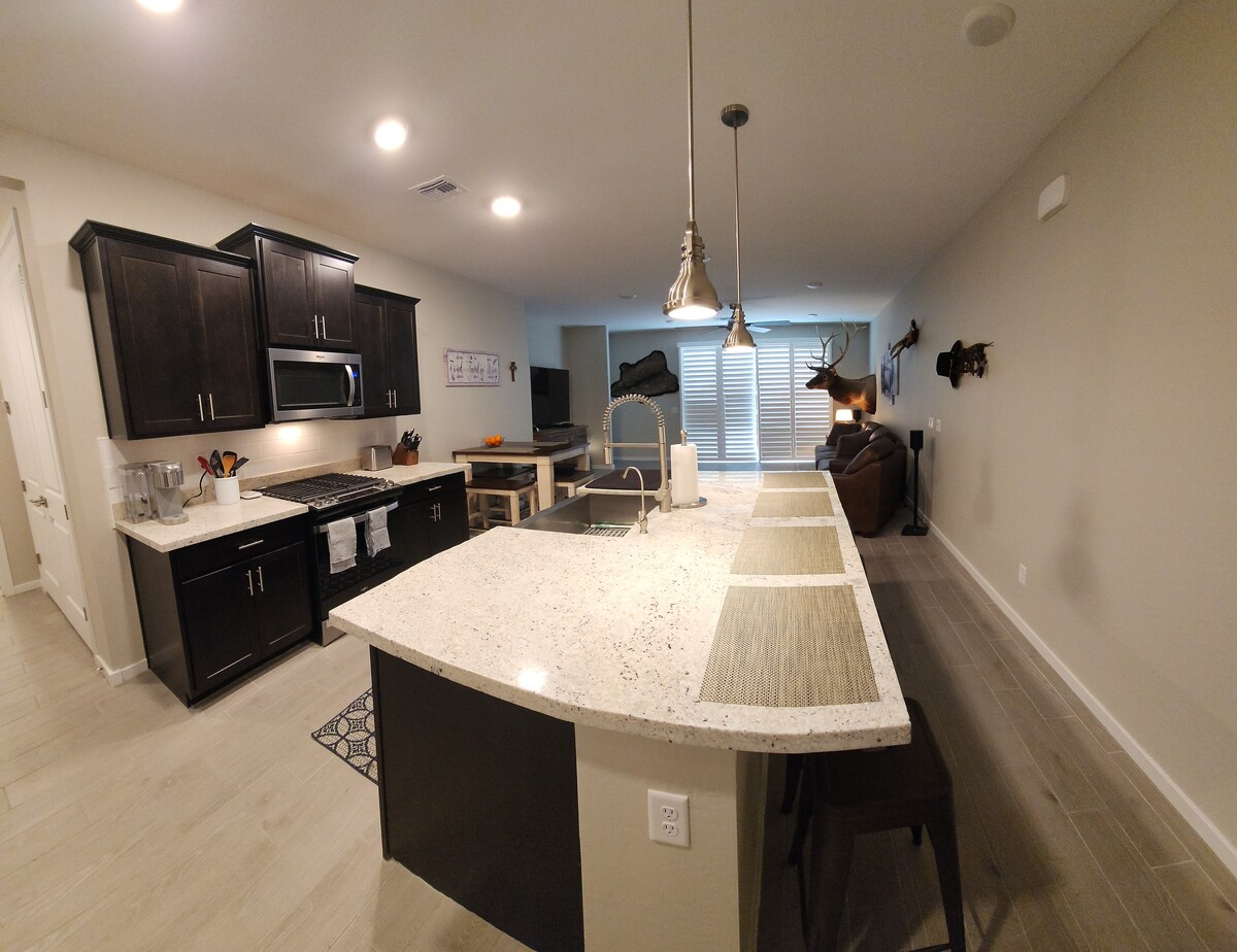 The kitchen features dark cabinetry and a large island with seating. A granite countertop is visible, along with modern appliances and a dining area in the background. Soft lighting illuminates the open space, providing a warm atmosphere. The floor is lined with large tiles.