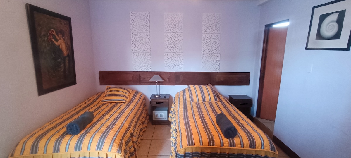 The room features two twin beds with yellow and blue striped bedding, complemented by matching towels. A bedside table with a lamp is positioned between them. Artwork adorns the walls, and natural light enters through a nearby door.