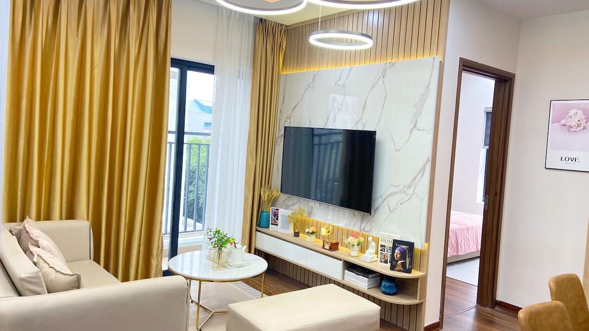 A modern living area features a light-colored sofa and a round coffee table. A large flat-screen TV is mounted on a marble wall, complemented by ambient lighting. Floor-to-ceiling curtains frame a window, allowing natural light to brighten the space, which has a warm, inviting feel.