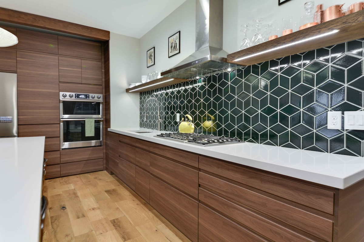 A modern kitchen features custom wooden cabinetry with sleek lines and a striking hexagonal tile backsplash in deep green. A built-in oven and microwave are visible, alongside a gas cooktop with a stylish hood. Natural light enhances the warm tones of the hardwood flooring.
