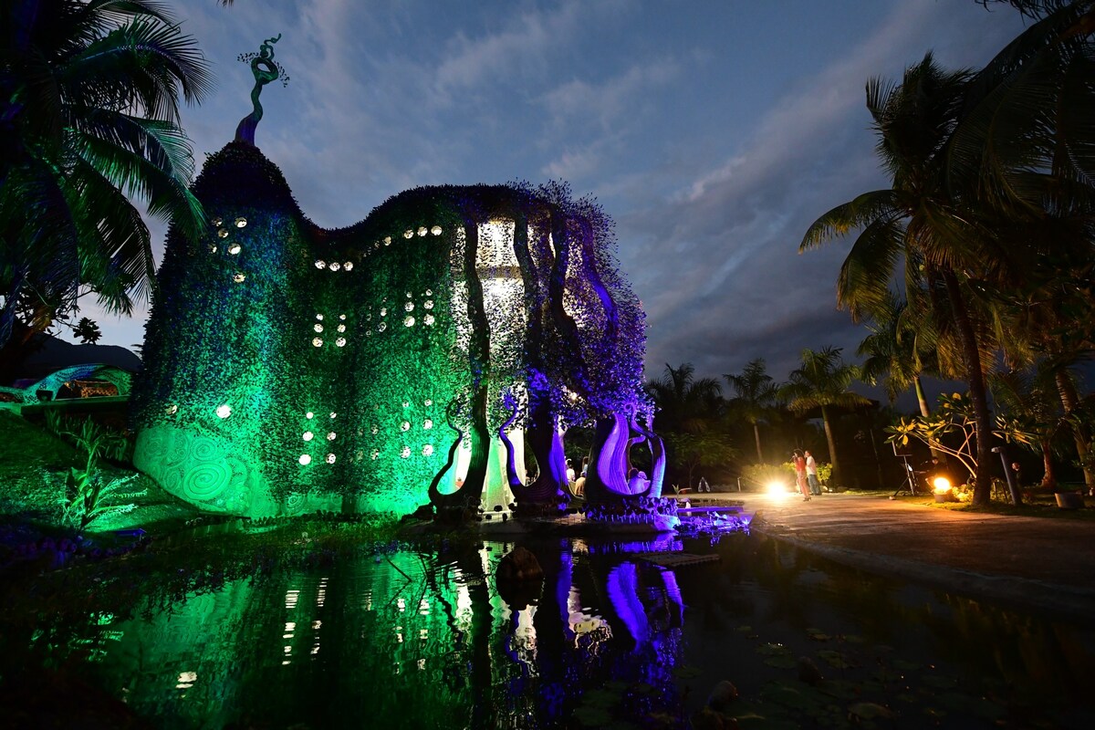 An artistic villa exterior is illuminated in green and purple lights, surrounded by palms. The building features unique architectural shapes resembling a peacock, with reflections visible on the water's surface in front. A pathway leads up to the structure, creating an inviting ambiance.
