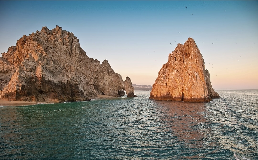 A serene coastal scene displays rugged rock formations emerging from gently lapping waves. The colors of the sky transition from warm hues to cooler tones, while the calm water reflects the landscape. Soft sands are visible along the shoreline, enhancing the natural beauty of the environment.
