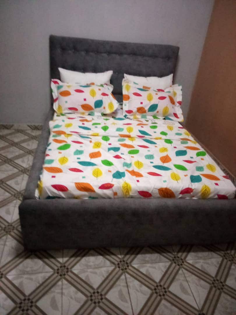 A spacious bed is dressed with a colorful leaf-patterned duvet cover and matching pillowcases. The bed's headboard has a soft gray upholstery, complementing the patterned bedding. The floor features a tile design with alternating geometric patterns in muted tones.