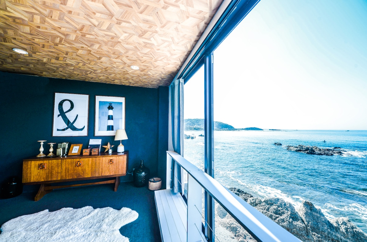 A spacious living area features large glass doors opening to stunning ocean views. Natural light enhances the wooden ceiling and walls adorned with nautical-themed artwork. A stylish console table holds decorative items, complementing the serene coastal atmosphere.
