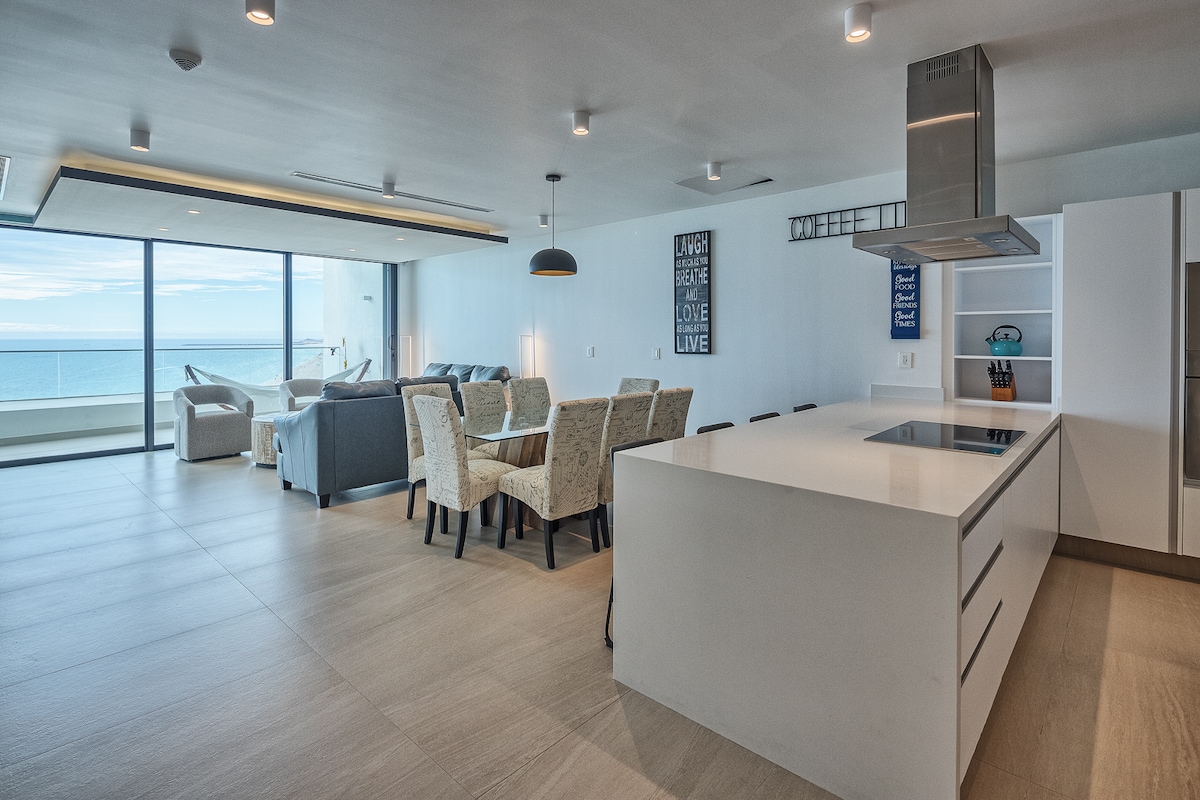 A modern open-concept living space offers a dining area with a wooden table surrounded by upholstered chairs. A kitchen island with a cooktop is featured in the foreground. Large windows showcase ocean views, allowing ample natural light into the area.