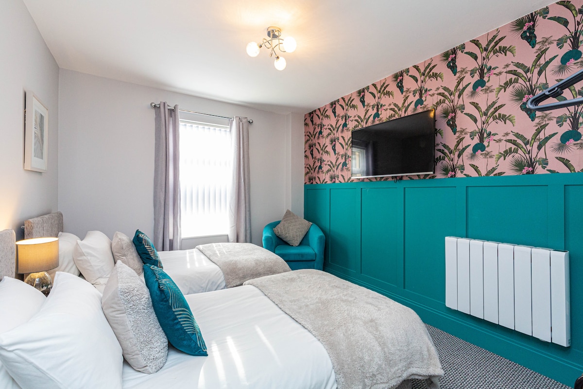 A tastefully decorated bedroom features twin beds with soft bedding, complemented by a stylish teal accent wall and tropical patterned wallpaper. A modern wall-mounted television is positioned at the foot of the beds, while a cozy armchair is placed in the corner, adding to the room's inviting design.