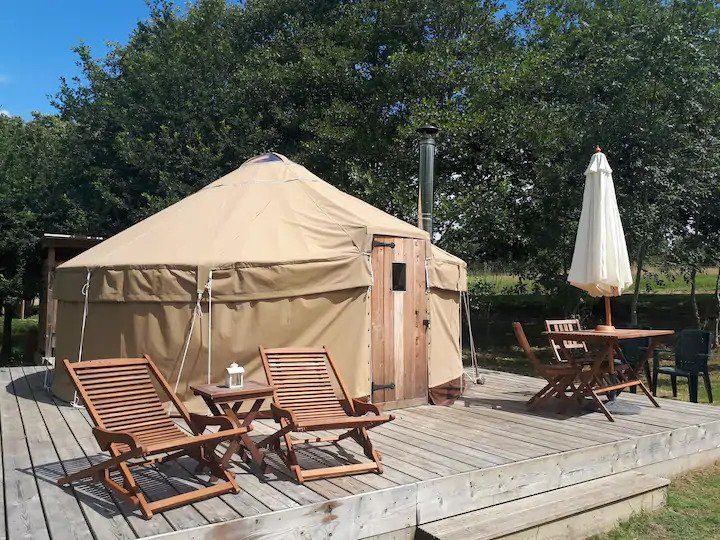 A spacious yurt is set on a wooden deck, surrounded by greenery. Two lounge chairs are positioned nearby with a small table between them. A dining table is visible, accompanied by a white umbrella providing shade. The entrance of the yurt is displayed prominently.