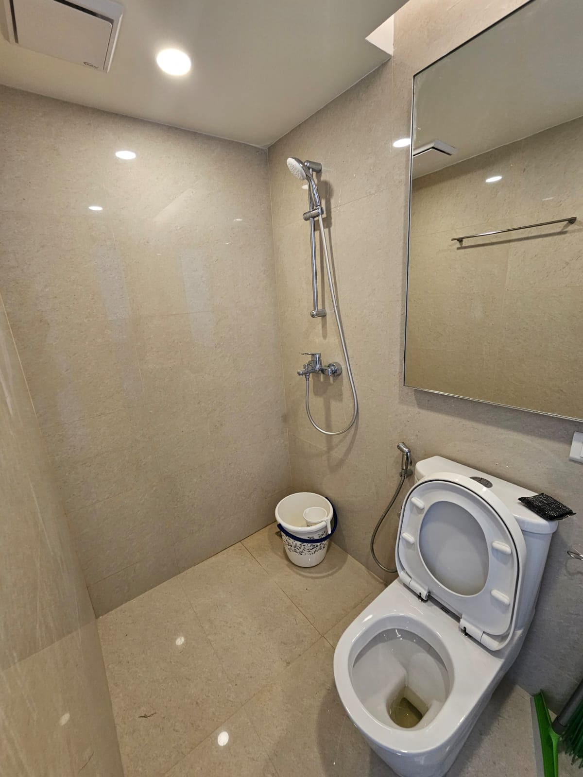The bathroom features modern fixtures, including a shower with a sleek chrome handheld head mounted on a tiled wall. A compact toilet is positioned next to a bucket placed on the floor. Soft lighting is provided by ceiling fixtures, enhancing the clean, minimalist design.