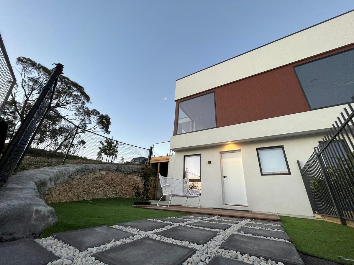 A modern building is presented, featuring large windows that allow natural light to fill the interior. The landscaped area includes a path of gray stones leading to an entrance. The surroundings showcase a grassy area and a decorative stone feature.