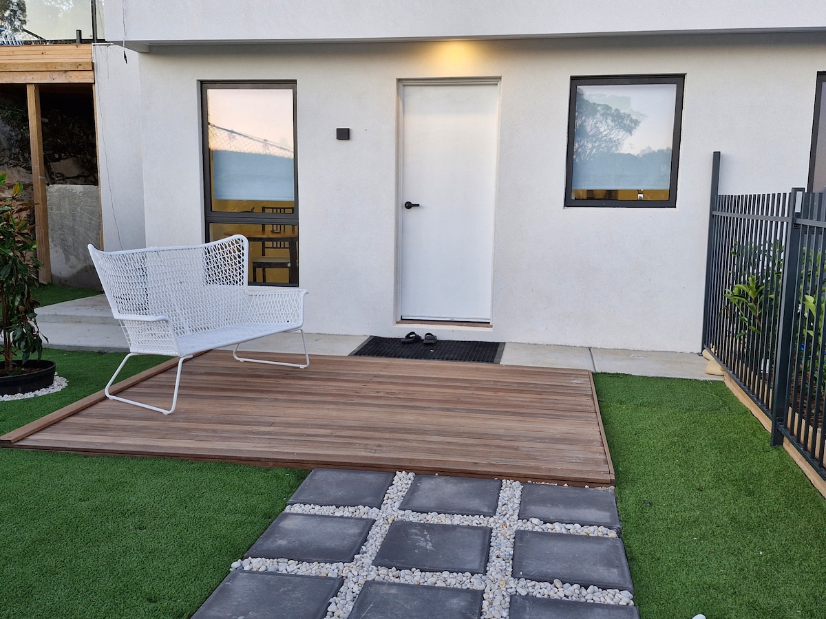 The entrance of the unit is shown, featuring a sleek white door flanked by two windows. A cozy seating area is positioned on a wooden deck, surrounded by well-maintained grass and decorative stones, offering a welcoming view of the surroundings.