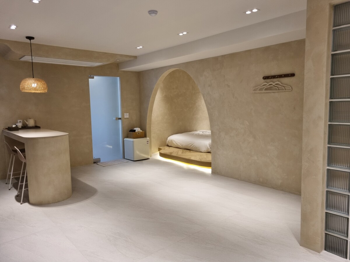 A spacious interior showcases soft beige walls and a minimalistic design. A cozy sleeping area is visible in an alcove, with a spacious bed positioned within it. A stylish kitchen island with stools adds functionality, and natural light enhances the tranquil atmosphere.