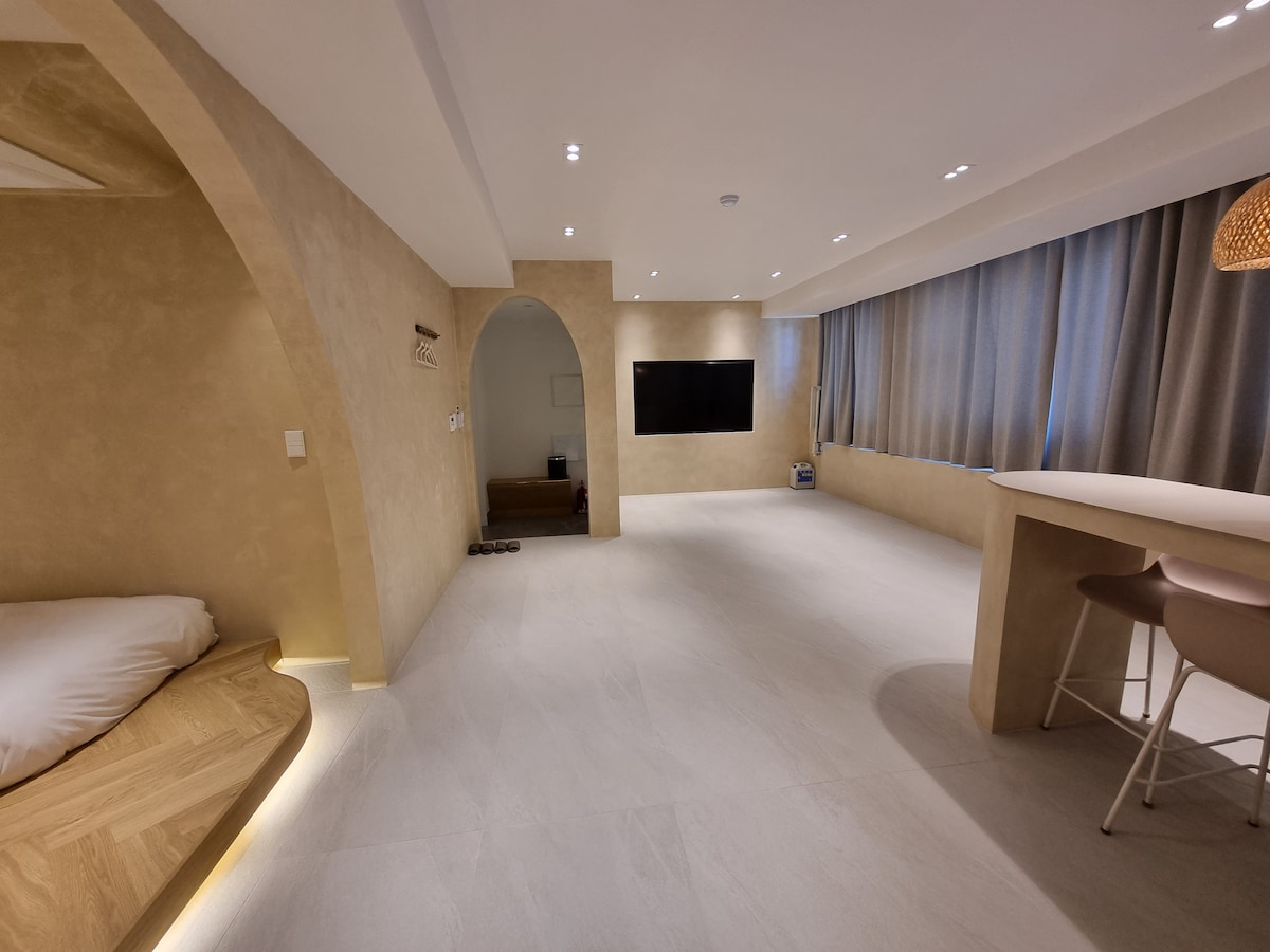 A spacious living area is showcased with light-colored walls and an open layout. A large wall-mounted TV is positioned opposite a curved alcove with a cozy sleeping area. Natural light filters through floor-to-ceiling curtains, enhancing the modern design.