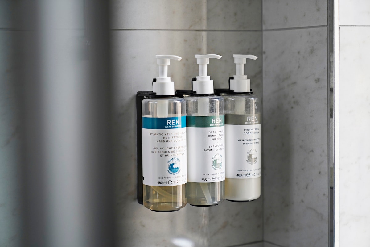 Three dispensers of personal care products are mounted on the wall in the modern shower. Each dispenser is labeled, providing easy identification of the shower gel, shampoo, and conditioner. The surrounding tiles are light gray, creating a clean and contemporary look.