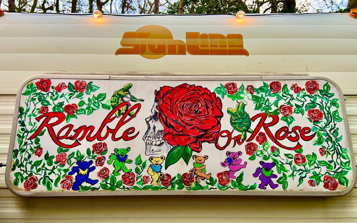 The camper's exterior features a painted sign displaying the name 'Ramble on Rose,' adorned with a large red rose, colorful flowers, and playful cartoon characters. The vibrant design reflects a cheerful atmosphere, set against the backdrop of the surrounding trees.