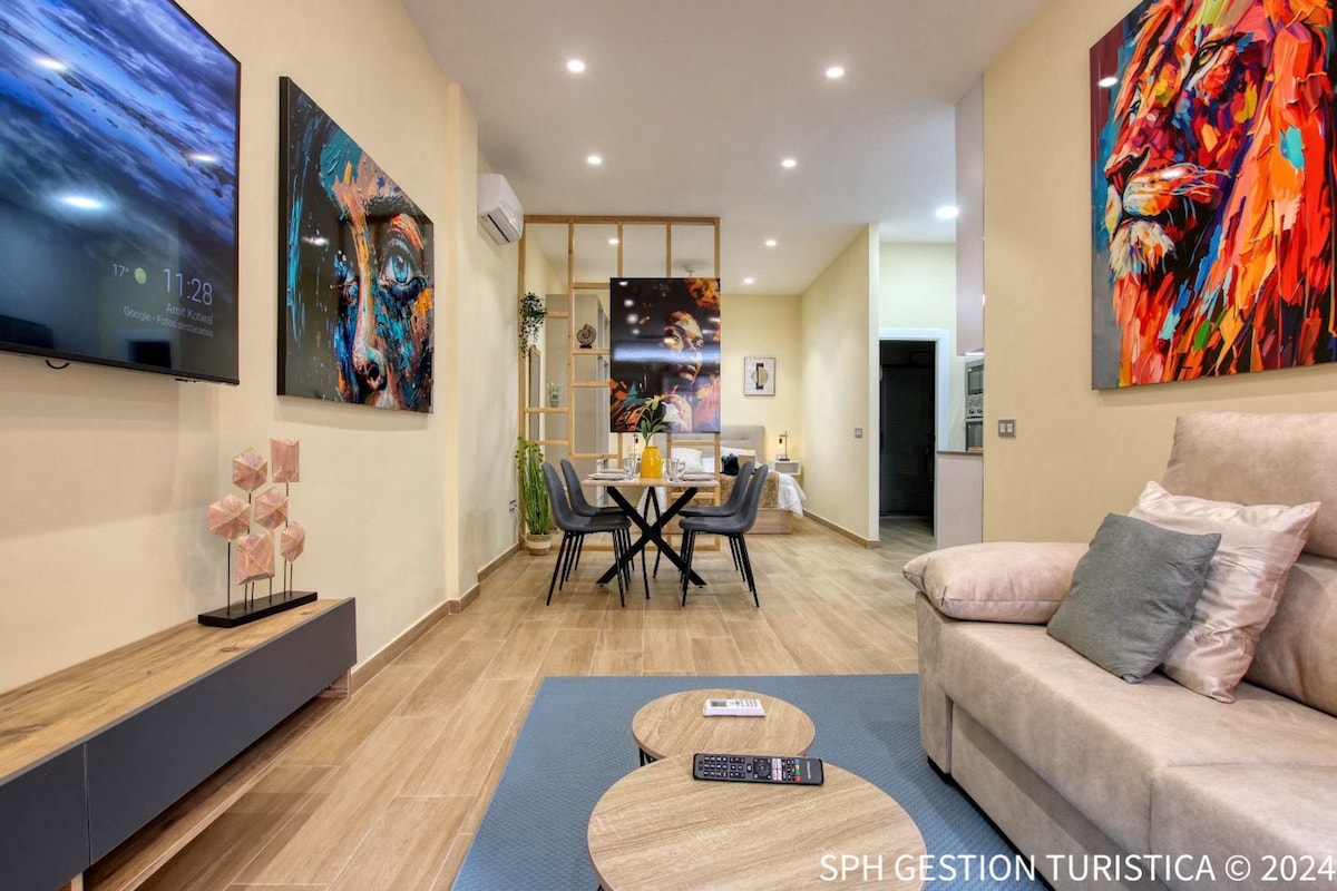 A modern living area features a comfortable sofa and a large flat-screen television mounted on the wall. Two vibrant artworks adorn the walls, while a round coffee table sits in front of the sofa, accompanying a dining table with four chairs in the background.