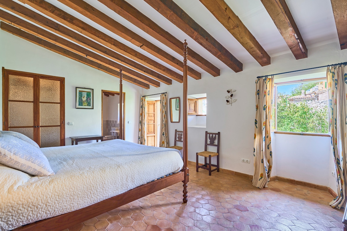 A bright and airy bedroom features a spacious wooden bed with a light-colored quilt. Exposed wooden beams line the ceiling, and natural light fills the room through multiple windows dressed with patterned curtains. A wooden chair and a small table add functional charm.