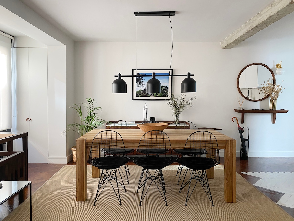 A dining area features a large wooden table surrounded by four black wire chairs. Three pendant lights hang above the table, providing focused illumination. A round mirror and a wall shelf with decorative elements are visible in the background, enhancing the modern design.
