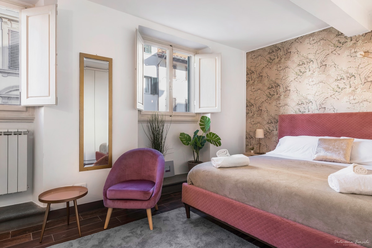 A comfortable bedroom featuring a spacious bed dressed in neutral tones. Two windows allow natural light to illuminate the space, complemented by a stylish armchair and a small table. A mirror reflects the room's design, enhancing the inviting atmosphere.