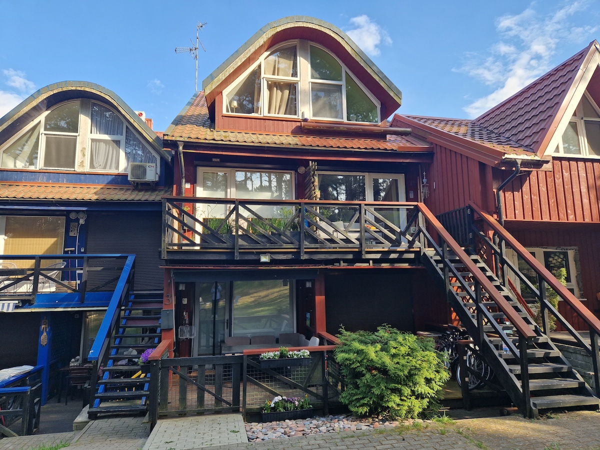 A two-story red wooden building features a steeply pitched roof and multiple large windows. A spacious deck is accessible by stairs, surrounded by greenery. The ground level includes a covered area, and bicycles are visible next to the structure, inviting outdoor activities.