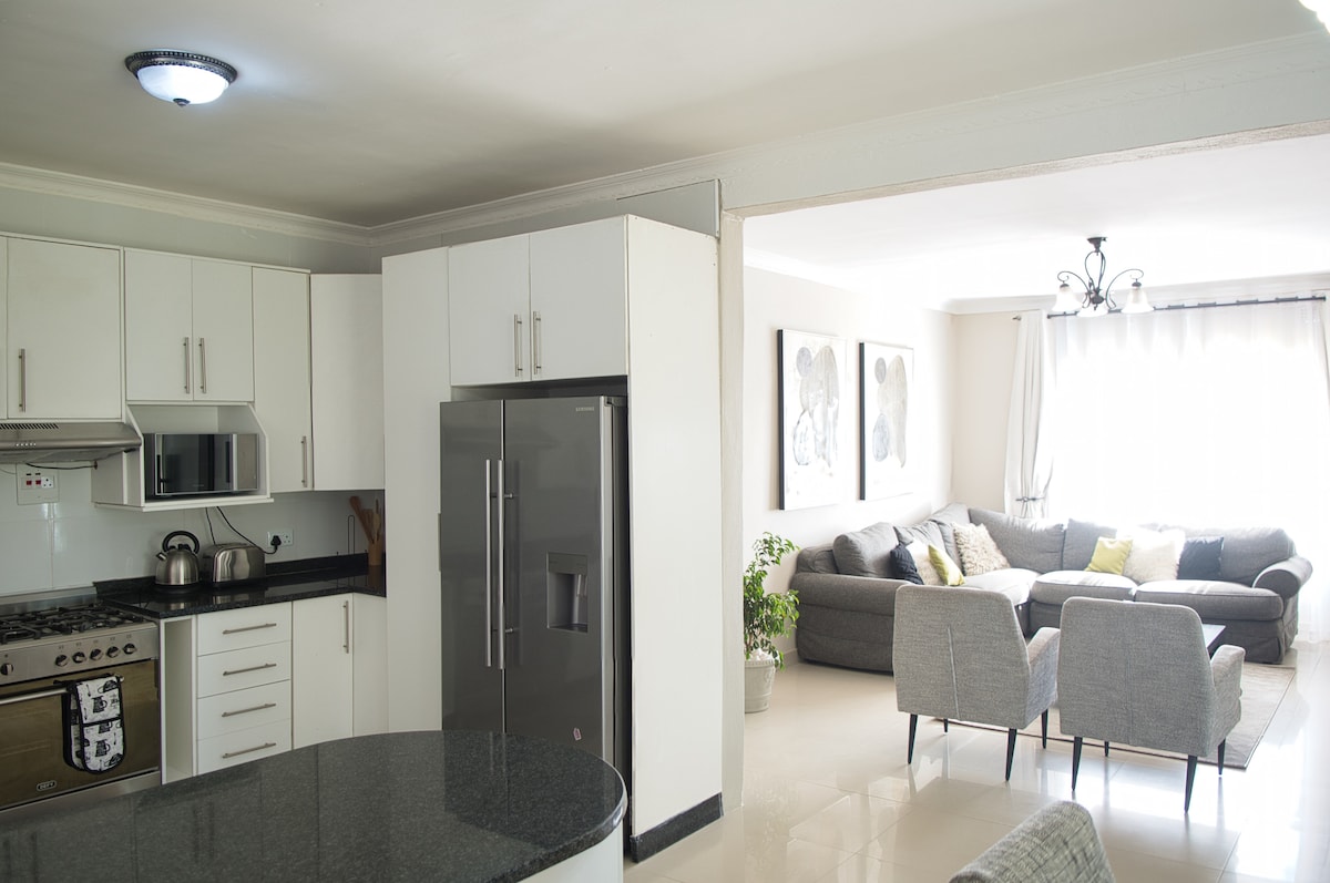 An open concept living area merges a modern kitchen and spacious lounge. The kitchen features sleek cabinets, a large refrigerator, and granite countertops. The living space, illuminated by natural light, includes a grey sectional sofa and two tasteful armchairs, with artwork adorning the wall.