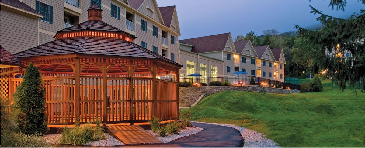 A wooden gazebo is set against a backdrop of a resort building, with warm lighting enhancing its structure. Lush green lawns surround the area, and multiple windows of the adjoining building reflect the evening light.