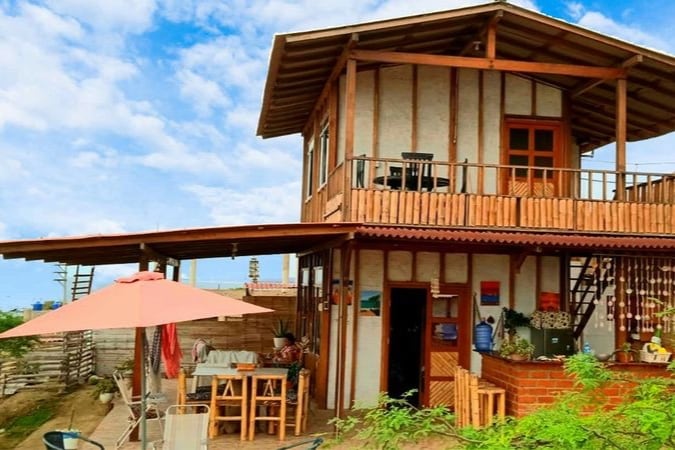 A two-story cabin features a wooden balcony with outdoor seating and a shaded umbrella. The entrance showcases a vibrant facade with large windows, surrounded by greenery and open space, enhancing the connection with the natural surroundings.