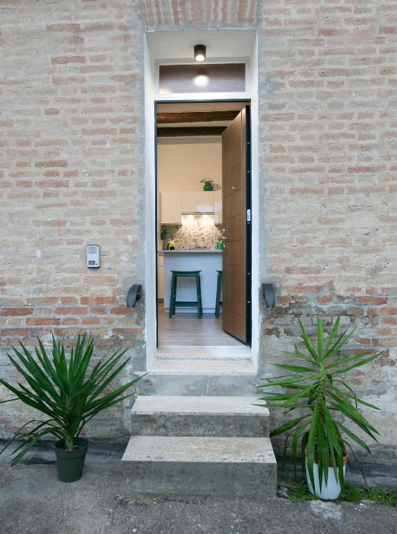 The entrance showcases a wide door leading into the apartment, with steps that guide the way. Potted green plants are placed on either side, complementing the rustic brick exterior. Light emanates from above, illuminating the entryway.