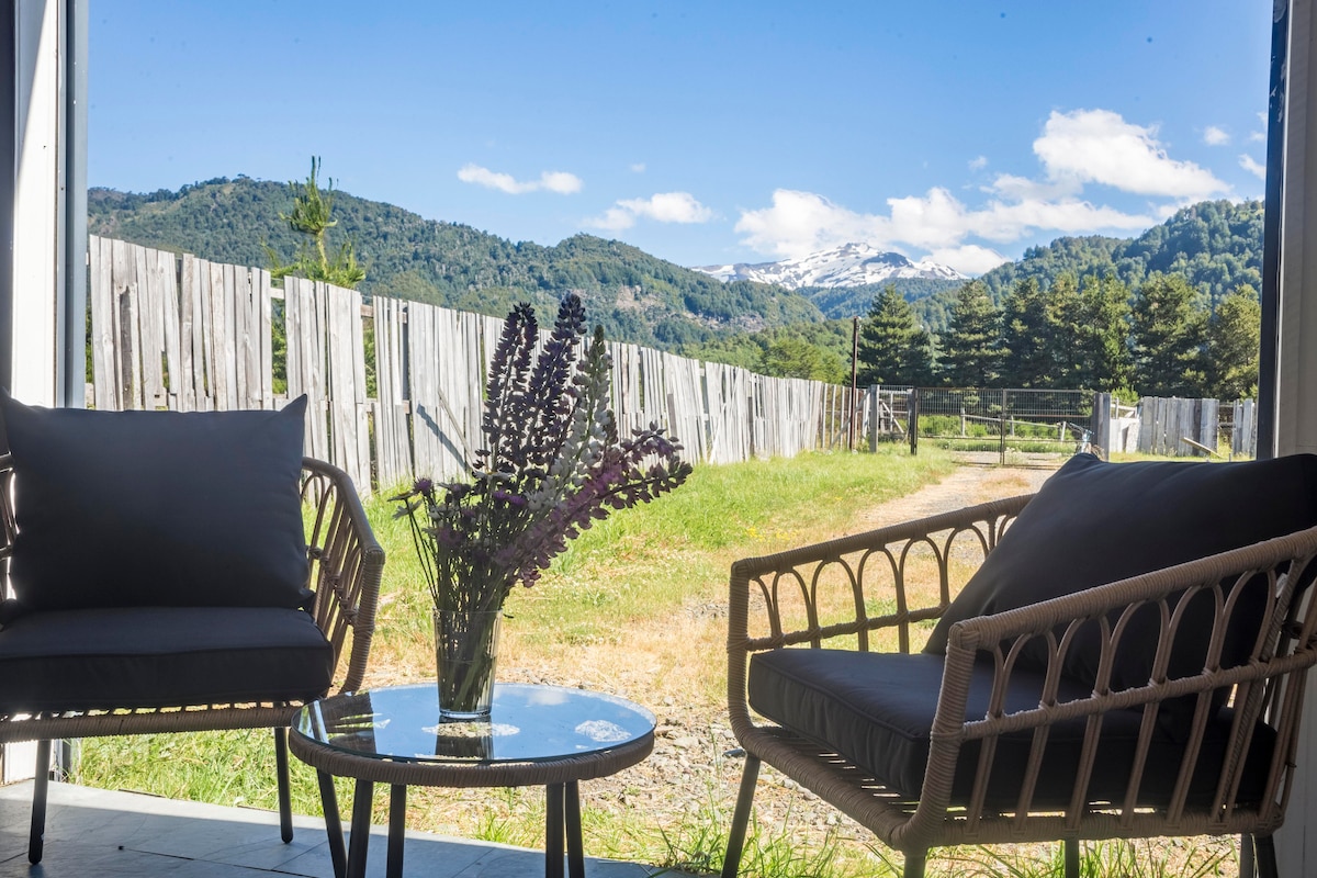 A serene outdoor seating area features two comfortable chairs and a glass table, with a vase of fresh flowers in the center. In the background, rolling hills and distant snowy peaks create a vibrant natural landscape under a clear sky.