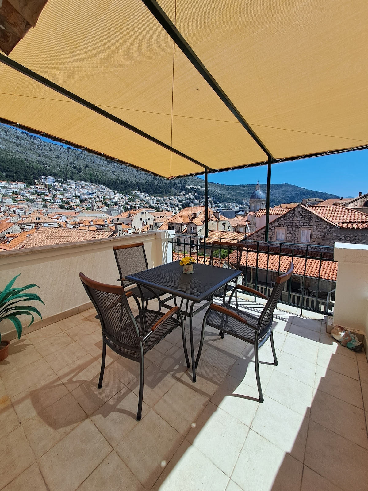 A spacious terrace is presented with a sturdy table surrounded by four chairs, shaded by a fabric canopy. A vibrant view of the city rooftops and surrounding hills can be seen in the background, showcasing the charm of the Old Town.