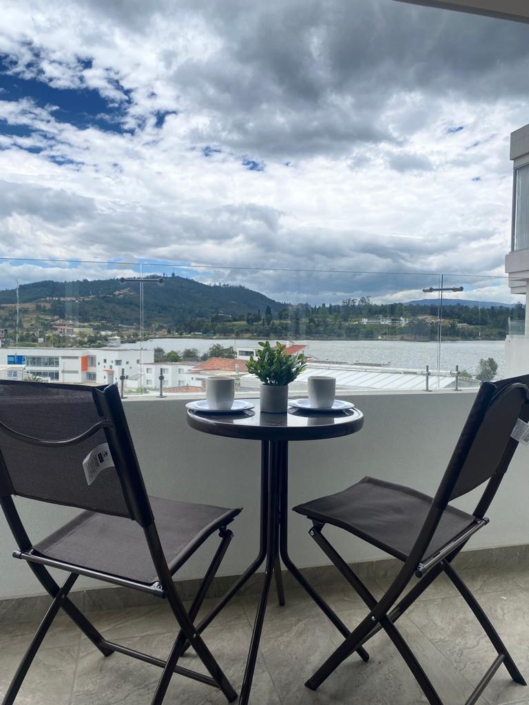A small balcony area is shown with a round table and two folding chairs. Centered on the table is a potted plant, with two cups placed on either side. In the background, panoramic views of hills and clouds are visible.