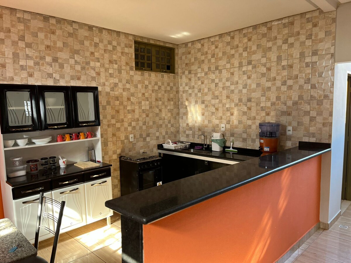 A modern kitchen area features a black granite countertop and sleek white cabinets. The wall is tiled with an earthy stone pattern. A stovetop and oven are neatly arranged, while various kitchenware is displayed on shelves. Natural light enters from a small window, brightening the space.