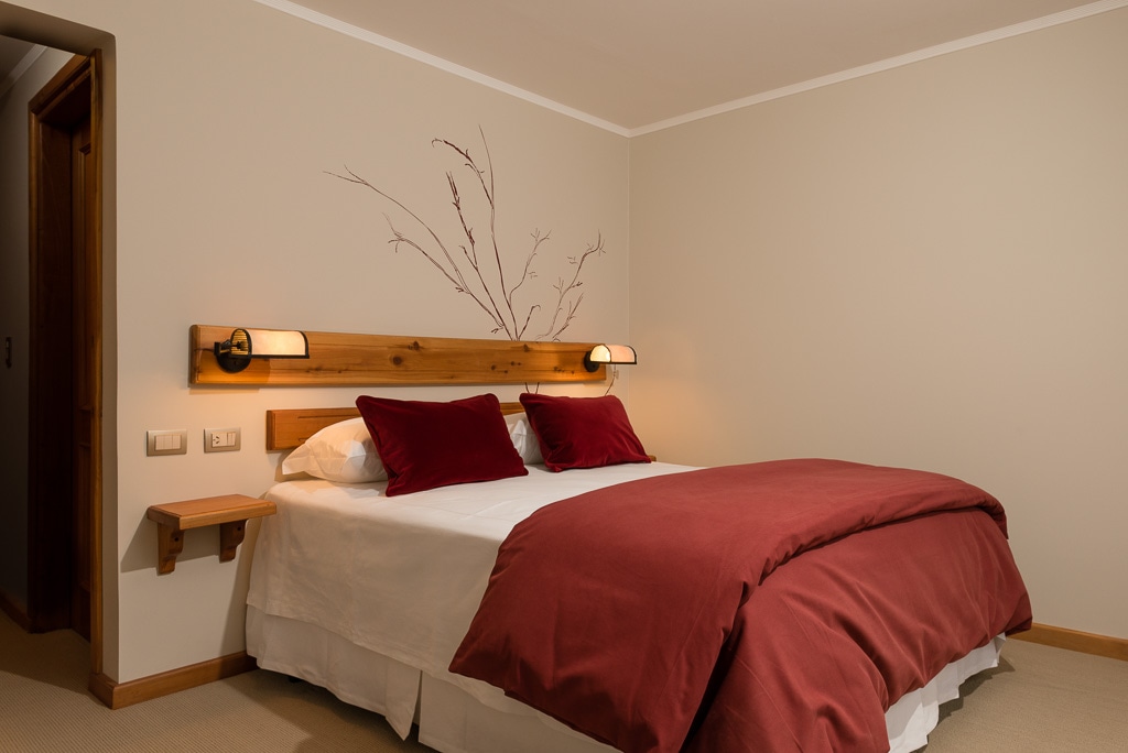 A cozy bedroom features a spacious bed with a deep red comforter and matching pillows. Wooden bedside lights are mounted above the bed, and a simple branch design is displayed on the wall. A small shelf is positioned beside the bed for convenience.