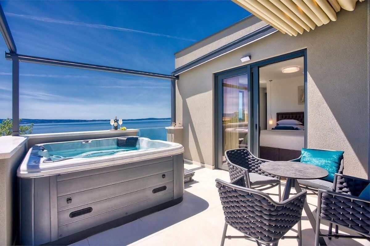 A private terrace is shown with a jacuzzi and a dining area, featuring a round table and four woven chairs. The backdrop includes expansive views of the sea, with clear skies and sunlight illuminating the space.