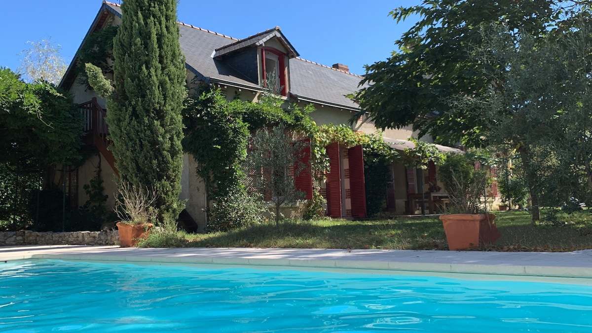 A tranquil view of the property is featured from the pool area, showcasing a charming building surrounded by greenery. The spacious outdoor space includes trees and planters, providing a natural ambiance and inviting atmosphere. The pool's clear water reflects the surroundings under a clear blue sky.