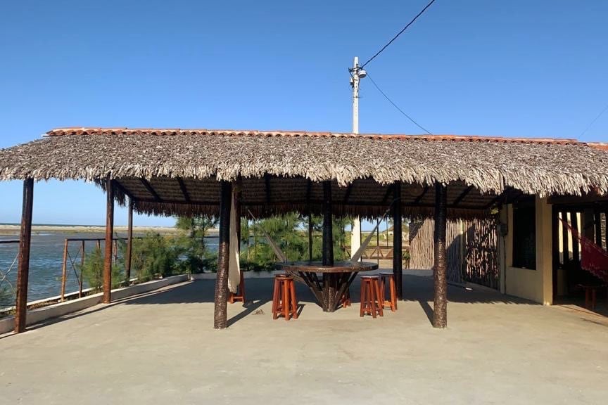 A spacious outdoor seating area is covered by a thatched roof, providing shade. Wooden tables and stools surround a large circular table, offering a setting for outdoor gatherings. Lush greenery and water views are visible in the background, creating a serene environment.