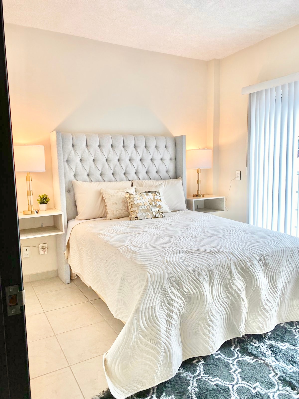 A contemporary bedroom features a plush tufted headboard with bedside tables and lamps on either side. A neatly made bed with a textured white quilt is complemented by decorative pillows. Natural light enters through vertical blinds, enhancing the serene atmosphere.